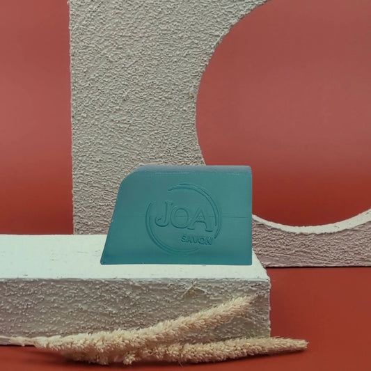 Savon solide Vétiver Bergamote de Joa, bleu-vert, posé sur socle blanc avec fond rouge texturé

